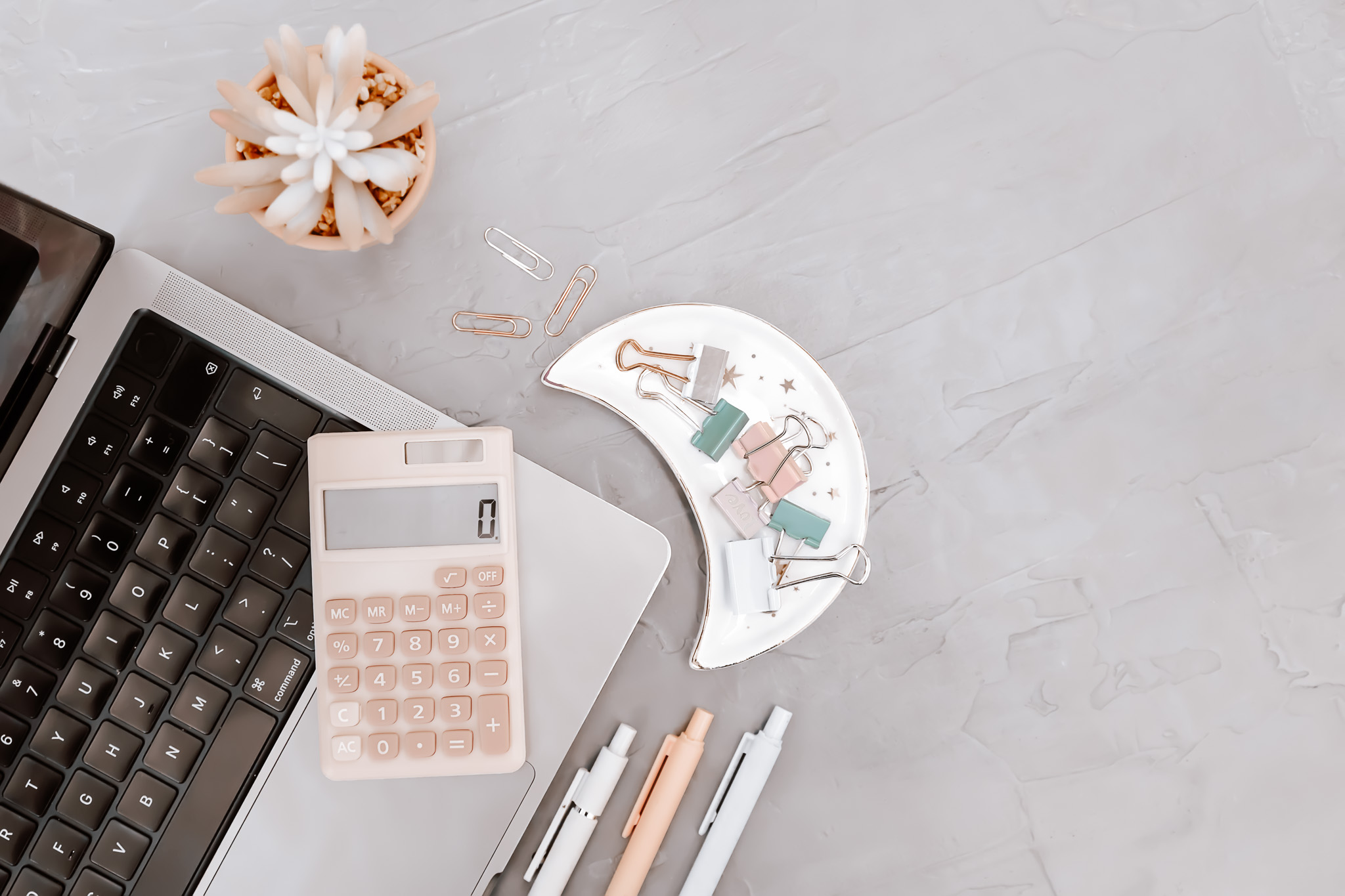 A pink calculator on top of a grey laptop next to 3 pens and a moon-shaped dish with paperclips